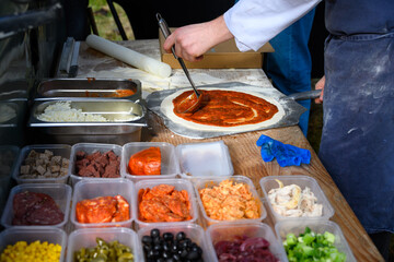 Chef ladling red sauce onto pizza dough, making a fresh pizza to cook in a wood fired oven outside in a food truck, lunch in the wild, Scotland
