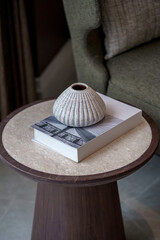 Cozy living room decor. A textured ceramic vase sits on a book, placed on a round wood and marble side table. Blurred armchair in background.