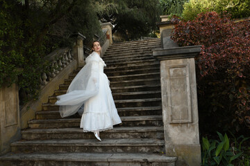Fototapeta premium portrait of beautiful young blonde model wearing historical white wedding gown and fantasy cloak. wandering ethereal figure walking away on historical staircase of fairytale castle location.