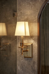 Elegant brass wall sconce light with a fabric shade, glowing warmly on a gray marble wall. Classic interior design detail for a luxury home.