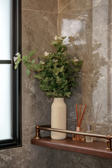 Elegant bathroom decor. Faux foliage with white blossoms in a ceramic vase, diffuser, and toiletries on a wood shelf against a gray marble wall.