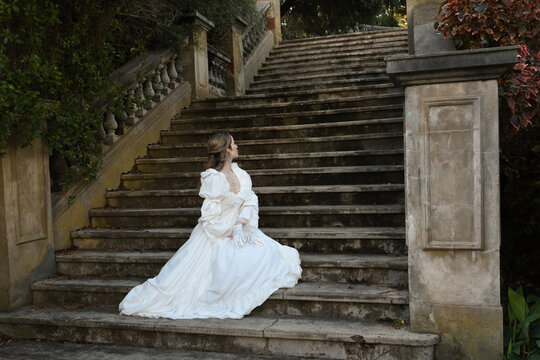 back view figure of female blonde model wearing white fantasy wedding gown. romantic sitting pose looking away on staircase of historical fairytale castle location 
