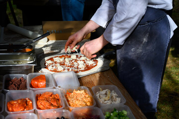 Chef placing fresh cheese on rolled out pizza dough on paddle to make individual wood fired pizza in food truck outdoors
