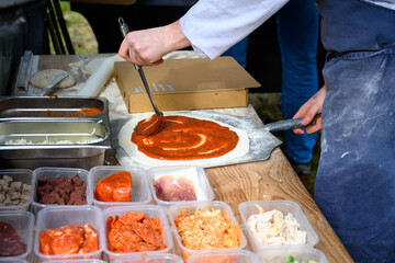 Chef ladling red sauce onto pizza dough, making a fresh pizza to cook in a wood fired oven outside in a food truck, lunch in the wild, Scotland

