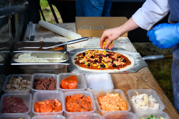 Chef placing fresh toppings on rolled out pizza dough on paddle to make individual wood fired pizza in food truck outdoors
