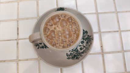 A glass of traditional teh tarik (pulled tea) with a distinctive layer of foam, served at a restaurant table in the evening. This warm beverage is popular in Southeast Asian eateries.