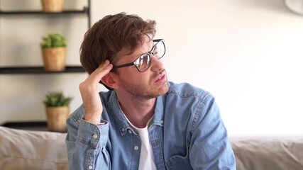 Tired young man sitting on sofa at home with hands on head, suffering from strong headache and stress, showing pain and exhaustion caused by fatigue, overwork, or mental tension indoors.
