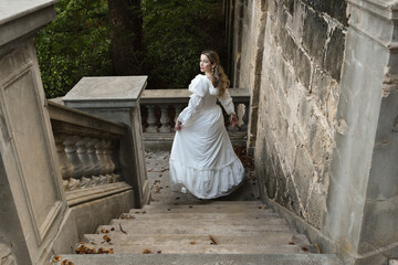 full length portrait of beautiful blonde female model wearing historical white gothic wedding gown, walking away back view, down staircase of romantic fairytale castle location with stone balconies