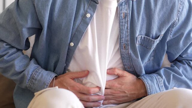 Young man sitting on sofa holding his stomach, feeling unwell and suffering from abdominal pain or digestive discomfort, showing sickness, nausea, and tension in a cozy home environment.
