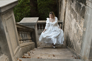 Naklejka premium full length portrait of beautiful blonde female model wearing historical white gothic wedding gown, walking down staircase of romantic fairytale castle location with stone balconies