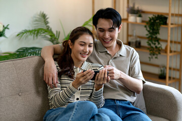 Asian young adult couple sitting on sofa happily watching smartphone enjoying digital content together home lifestyle