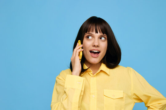 A cheerful young woman wearing a bright yellow shirt speaks on a mobile phone, smiling with surprise against a vivid blue backdrop, capturing a candid moment of friendly communication and modern