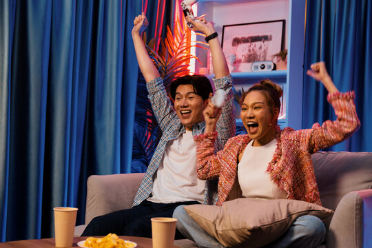 Couple gamer raising fist up holding joysticks be winner together complete level sitting sofa playing video game with front snack and drinks at neon light color living room at modern home. Infobahn. - Powered by Adobe