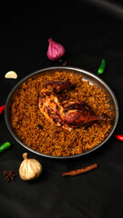 Roasted Chicken with Spiced Rice in Metal Dish Surrounded by Garlic Red Onion Green Chili Cinnamon Star Anise Black Peppercorns on Black Cloth
