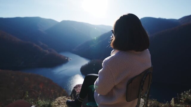 A static medium shot from behind a woman wearing a backpack, sitting on a cliff edge, drinking from a mug, and gazing at a beautiful mountain and river landscape. Features a bright sun flare.