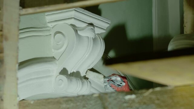 Extreme close-up shot showing a worker's gloved hand carefully painting a detailed, scroll-shaped, white plaster corbel or console bracket during an interior restoration project.