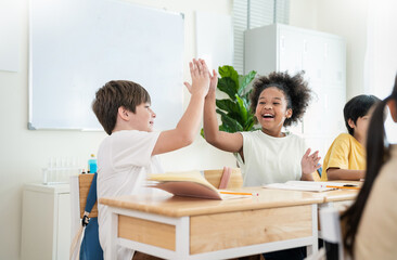 Two cheerful students caucasian and black sit at desks in classroom, smiling giving high five while studying together with notebooks open. Education back to school
