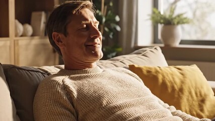Relaxed Man in Cream Sweater Resting on Gray Sofa with Yellow Pillow in Sunlit Room Near Window Peacefully Smiling In a Indoor Setting