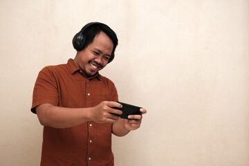 Southeast Asian man in a brown shirt smiling while playing a mobile game on his smartphone, wearing headphones in an indoor setting.
