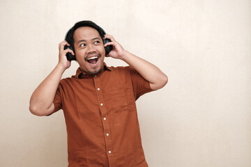 Southeast Asian man in a brown shirt looking joyful and surprised while listening to music on...