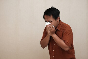 Southeast Asian man in a brown shirt sneezing or blowing his nose into a tissue, showing signs of illness or allergy indoors.