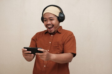 Smiling Southeast Asian man wearing headphones and beanie, enthusiastically playing a mobile game...