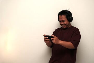 Smiling Southeast Asian man wearing headphones focused on playing a mobile game on his smartphone while standing indoors.
