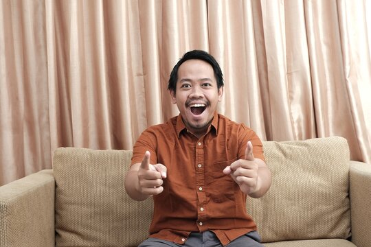 Smiling Southeast Asian man sitting on a sofa, enthusiastically pointing both fingers forward with a joyful and energetic expression.
