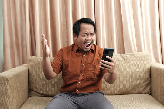 Frustrated Southeast Asian man sitting on a sofa, shouting at his smartphone with raised hand, expressing anger or disbelief indoors.
 - Powered by Adobe