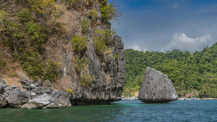 Picturesque cliffs in the ocean. Tropical green vegetation grows on the steep slopes. Boats off the coast of the island. Sandy beach. Blue sky, clouds. Philippines. Palawan. El Nido