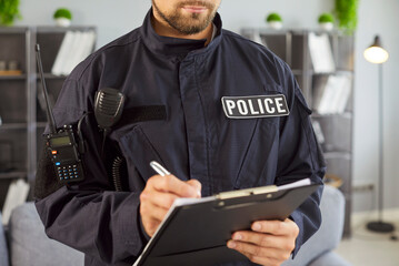 Close-up male police officer dressed in uniform stands indoors writing accident or criminal details...