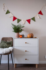 Fir branches and Christmas decorations in a vase, candle, pumpkin on white dresser. Christmas garland on wall. Brown chair with green pillow and white fur.