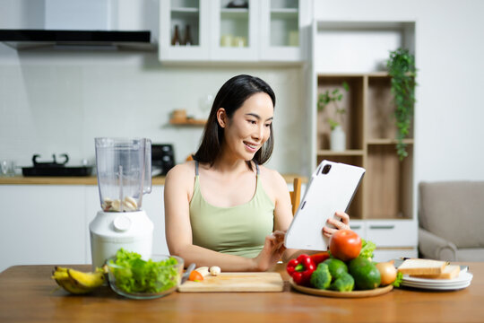 Smiling Asian woman using tablet surrounded by fresh vegetables in kitchen. Concept of healthy lifestyle, cooking, blogging, and digital wellness