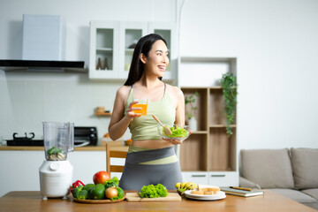 Smiling Asian woman in sportswear holding fresh salad and orange juice in kitchen. Concept of health, diet, wellness, and fitness lifestyle.