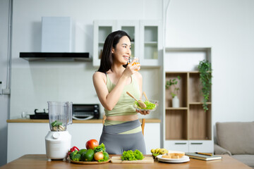Smiling Asian woman in sportswear holding fresh salad and orange juice in kitchen. Concept of health, diet, wellness, and fitness lifestyle.