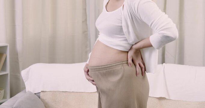 Asian expecting woman gently holds exposed pregnant belly while standing indoors reflecting maternal love, connection, preparation, and peaceful anticipation in calm third trimester lifestyle