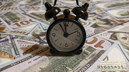 A black alarm clock placed on scattered US hundred-dollar bills, symbolizing the relationship between time and money, financial management, investment, and economic value.
