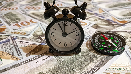 A black alarm clock and compass placed on a spread of hundred-dollar bills, symbolizing time, wealth, and guidance in financial decisions and investment strategies.