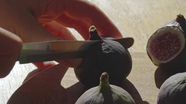 A hand carefully cuts fresh figs on a wooden board. The kitchen is bright and warm, showcasing the vibrant colors of the fruit. Perfect for summer recipes and healthy snacks.