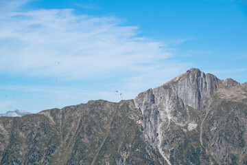 Ridge of brown-green mountains with numerous paragliders soaring in clear blue sky