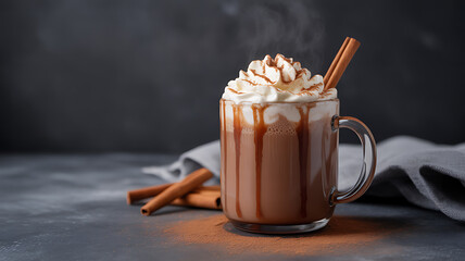 A glass mug filled with a caramel-drizzled hot beverage topped with whipped cream and a cinnamon stick, set on a dark surface with additional cinnamon sticks nearby.