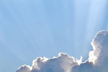 Bright crepuscular sun rays fan out across a clear, pale blue sky from behind a large, fluffy white cloud on the bottom edge of the frame, providing ample copy space.