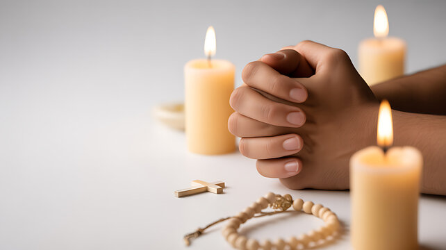 Hands clasped in prayer with lit candles, rosary, and cross on a white surface