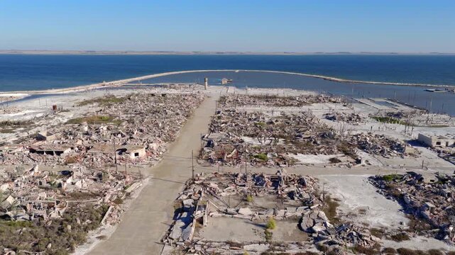 Drone camera frames the main avenue of Villa Epecu&eacute;n, shooting over rows of destroyed and salt-covered buildings, converging toward the old pier and the expansive blue waters of the lake.