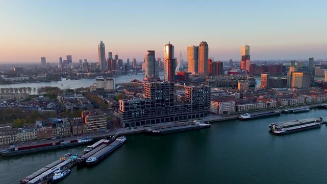 Aerial drone footage of Rotterdam Netherlands showing modern riverfront skyline, Erasmusbrug district, and illuminated buildings reflecting warm sunset light over Maas River and harbor area