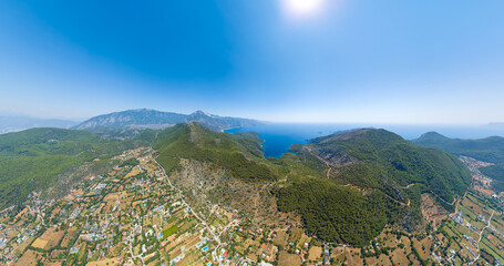 Kayakoy, Turkey. Aerial panorama showing modern town development alongside ancient ghost town ruins on forested mountain slopes with Mediterranean sea view under clear summer sky. Aerial view