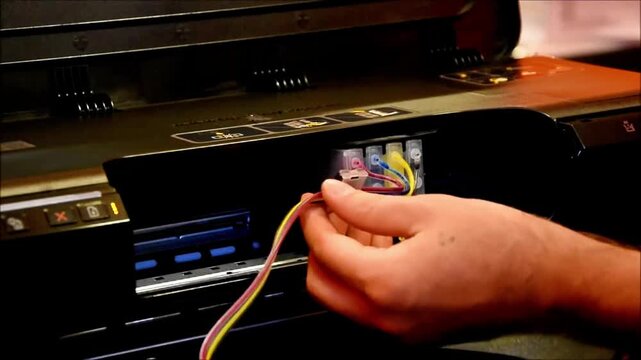 Technicians hands guide flexible ink tubes into a large-format printer system, aligning connectors and securing flow paths for smooth, high-volume color printing setup