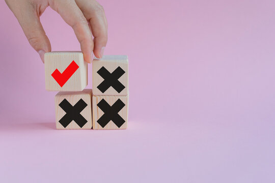 A hand places a wooden block with a red check mark above three blocks with black crosses. The background is a soft pink color, emphasizing the concept of choice and decision-making.
