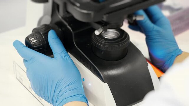 Close up of scientist hands in blue gloves adjusting microscope lenses in blood analysis laboratory indoors for medical diagnostics and research.