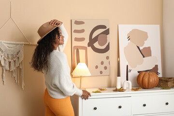 Beautiful young happy African-American woman in stylish outfit with autumn leaves, pumpkin and hat...